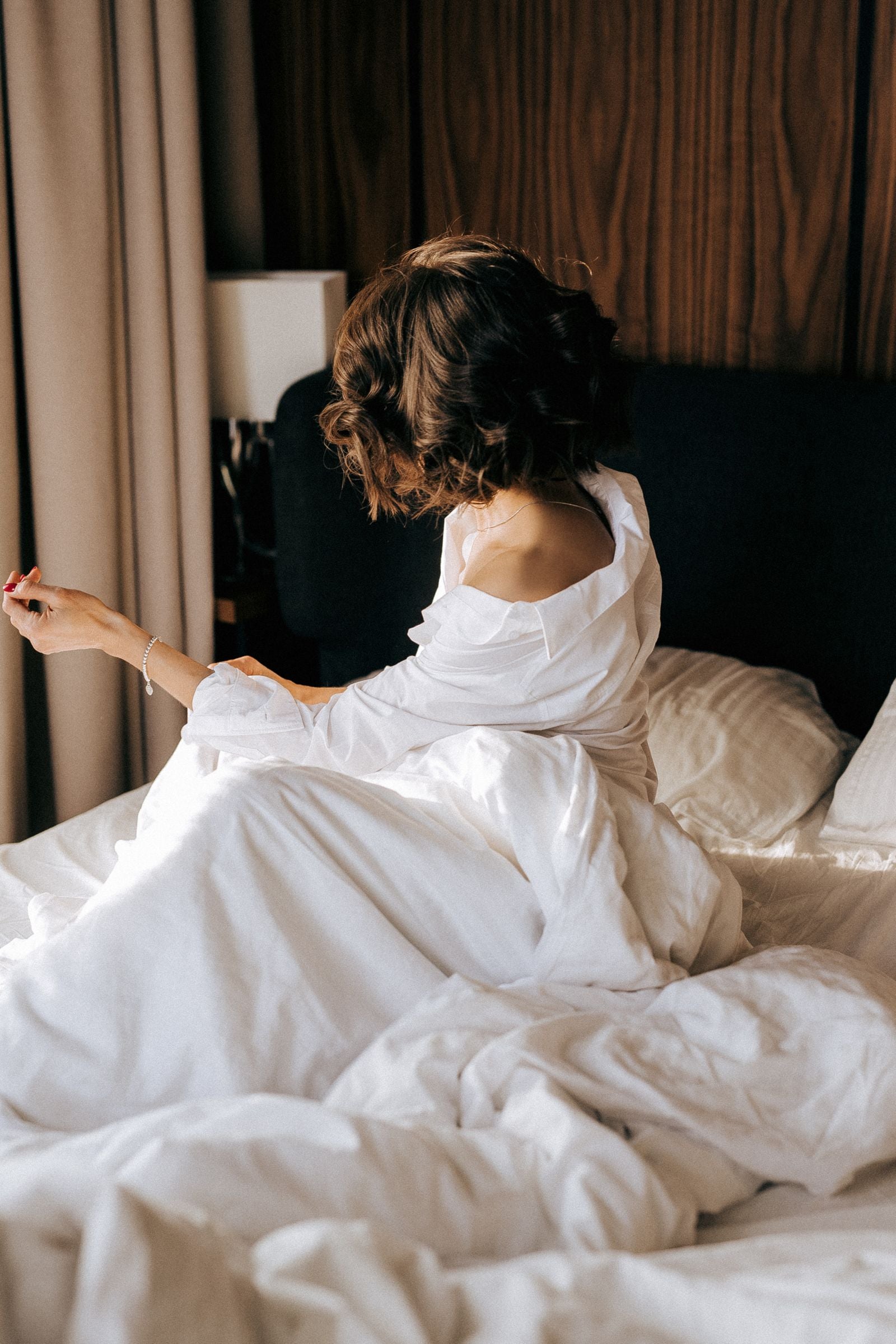 Woman in a soft white shirt slipping off one shoulder, sitting in morning bedlinen