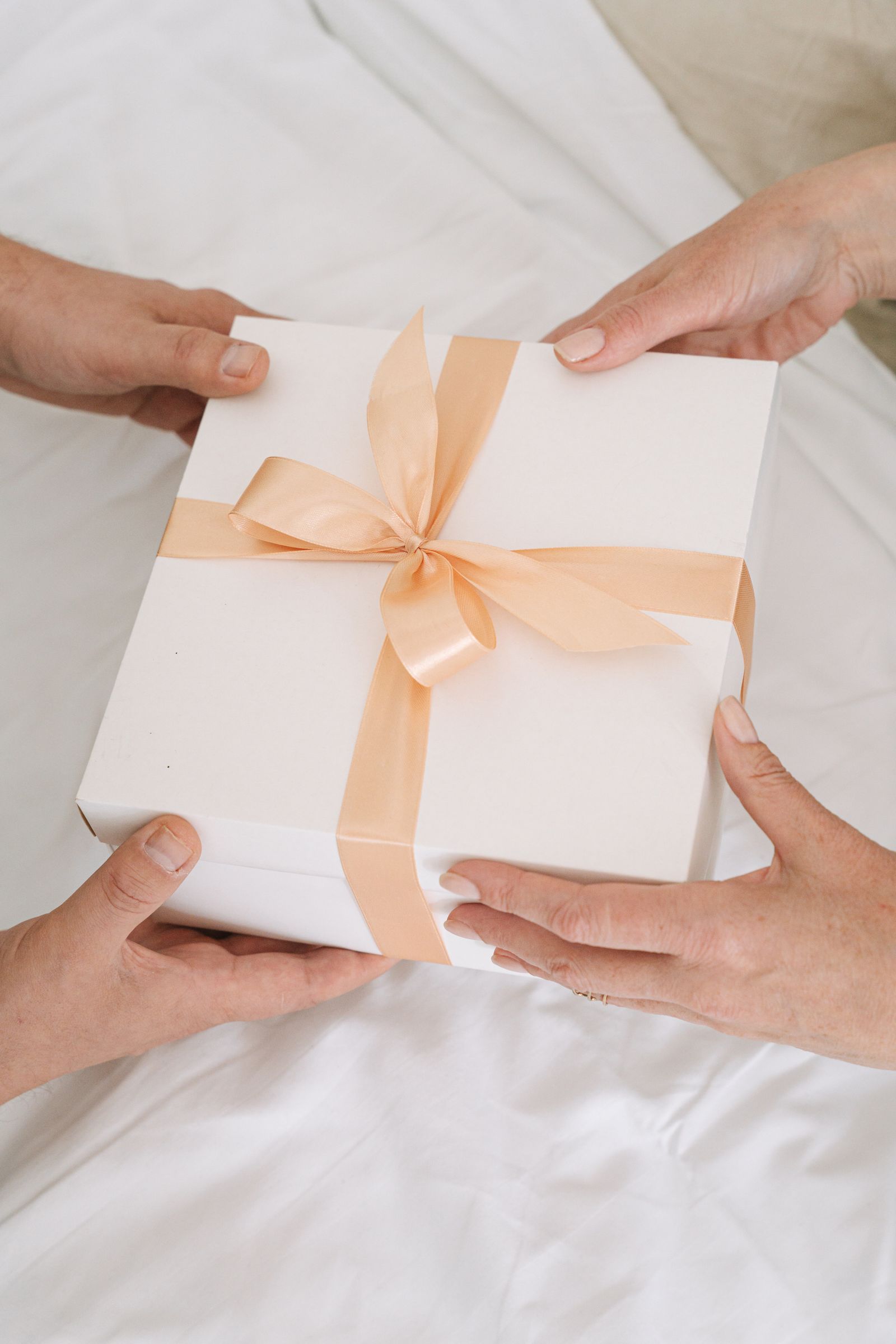Hands exchanging a white gift box tied with a peach satin ribbon on bedding